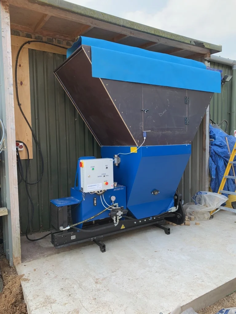 Falach 100 briquette machine in blue with a large hopper and control panel, installed inside a timber and metal shed.