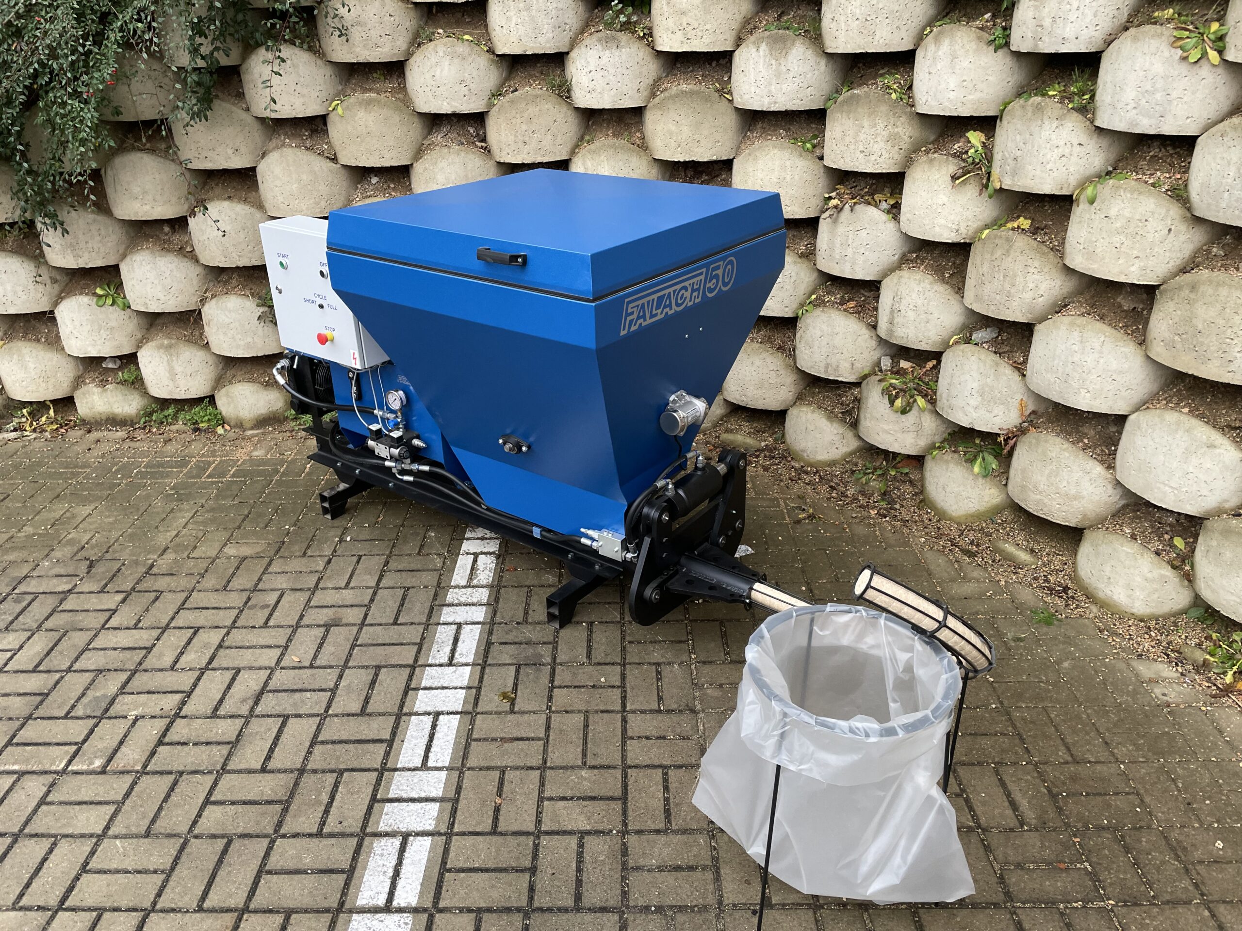 Falach 50 briquette machine in blue with control panel, displayed on a brick paved surface with a collection bag alongside